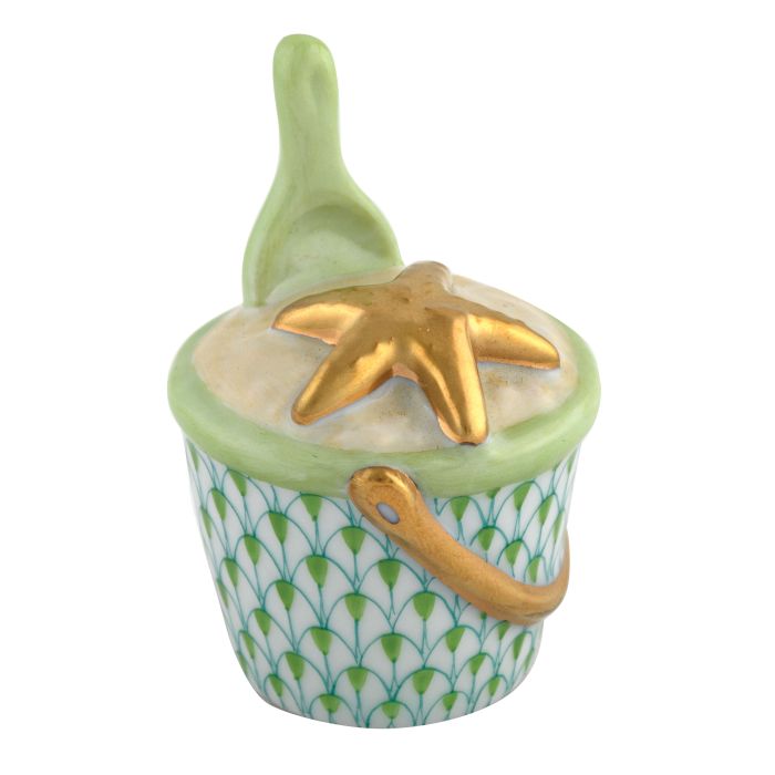 The Herend Beach Bucket Collection features a decorative white porcelain container shaped like a sand bucket with a green handle, green-and-white pattern, gold starfish lid, and a matching hand-painted ceramic spoon.