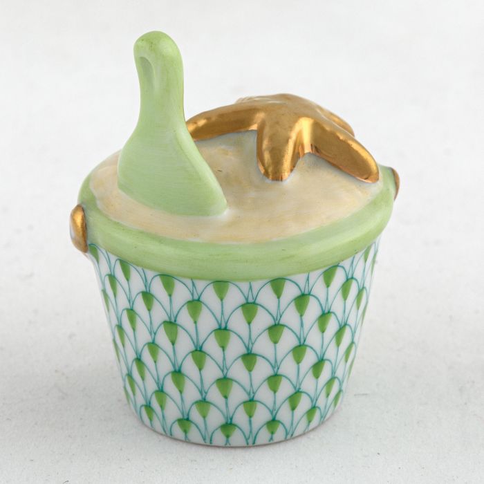 The Herend Beach Bucket Collection features a small decorative porcelain container with a green and gold fish scale pattern, a light green lid, and hand-painted gold sea star and shell accents on top.