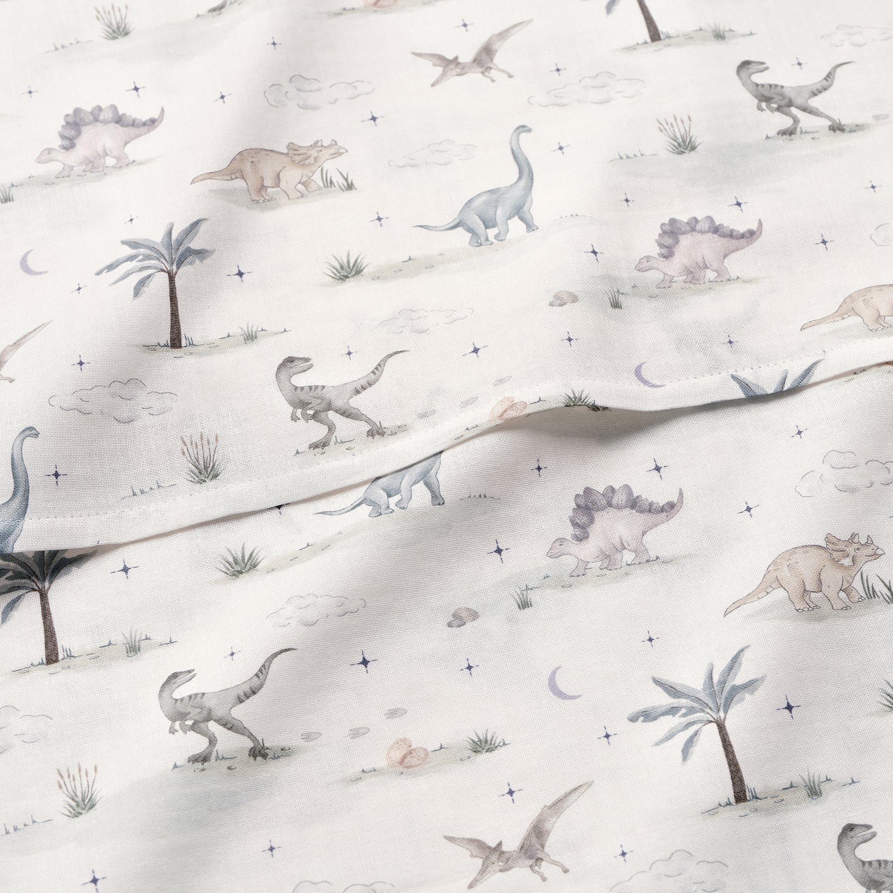 The Gooselings Dino Dreams Swaddle features soft pastel dinosaurs, palm trees, clouds, and stars on 100% white cotton—ideal as a cozy swaddle or baby blanket.