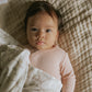 A baby with dark hair and a pink outfit lies on a textured beige blanket, partially covered by the Gooselings Animal Parade Ivory Swaddle, a cozy swaddle blanket made from 100% cotton.