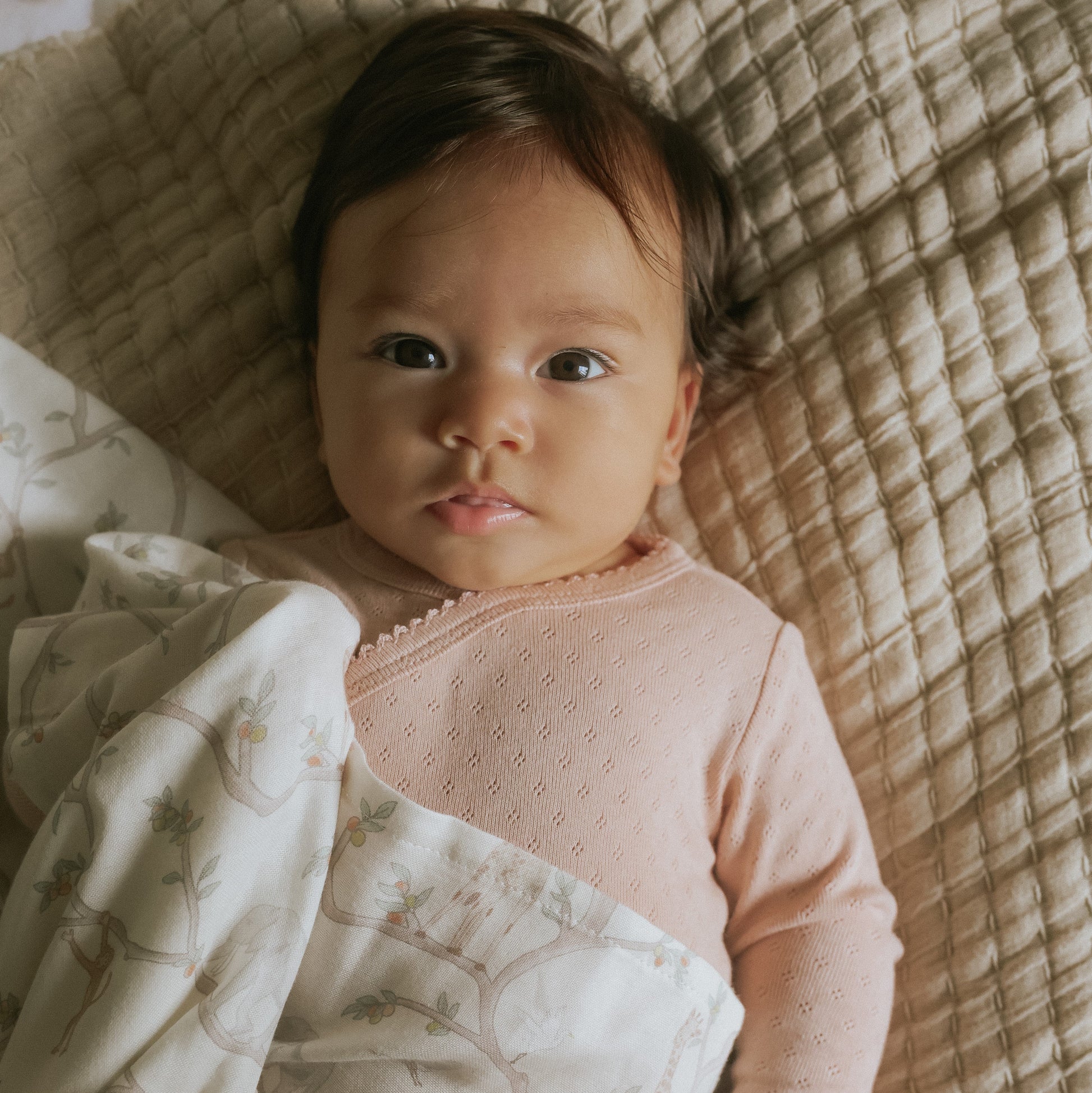 A baby with dark hair and a pink outfit lies on a textured beige blanket, partially covered by the Gooselings Animal Parade Ivory Swaddle, a cozy swaddle blanket made from 100% cotton.