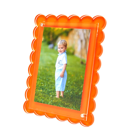 A framed photo of a child on green grass, showcased in Tara Wilson Designs’ best-selling Scallop Frame Collection—an orange, scalloped-edge lucite frame that adds brightness to any space.