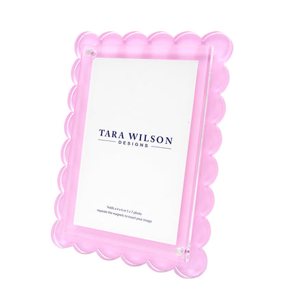 The Scallop Frame Collection by Tara Wilson Designs features a pink lucite scalloped frame that stands upright, stylishly displaying photos and adding a chic touch to any space.