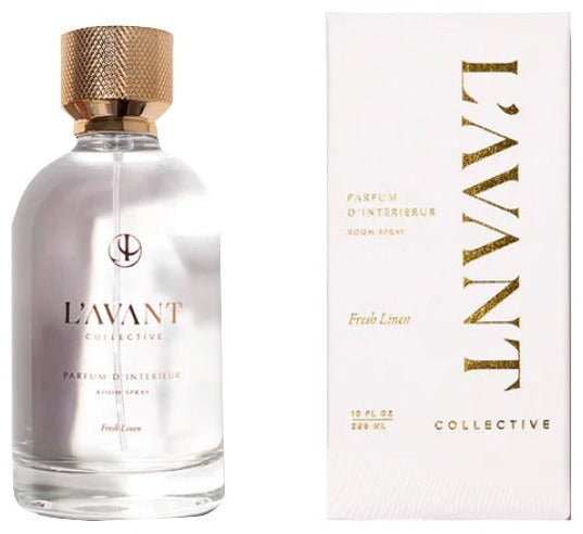 A clear glass bottle of L'Avant Collective Fresh Linen Scent Collection room spray is displayed next to its elegant white and gold box; the collection also includes hand soap and laundry detergent.