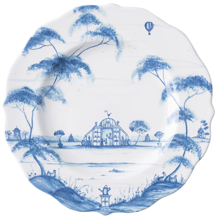The Juliska Country Estate Delft Blue Dinnerware Collection features a decorative porcelain plate with a blue pastoral scene—trees, central pavilion, small animals, and hot air balloon—on a scalloped white background.