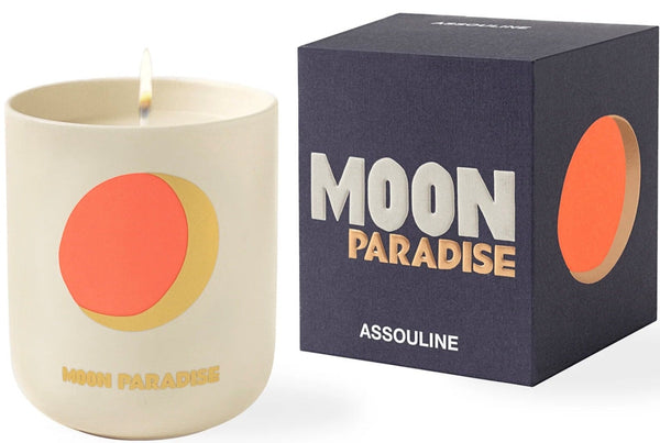 A lit candle with a stylized moon design next to its packaging box labeled "Travel Candle Moon Paradise - Assouline".