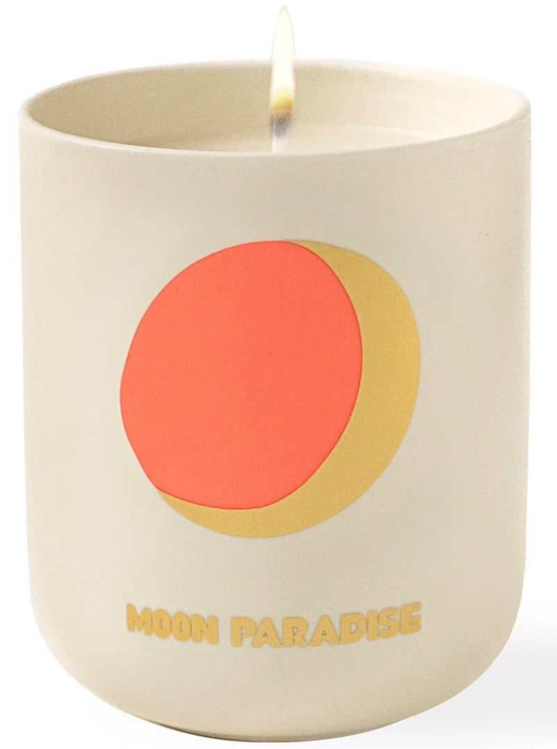 A lit scented candle with a minimalist design, featuring a red and gold circle motif and the words "Assouline Travel Candle Moon Paradise.