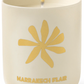 A lit Assouline Travel Candle Marrakech Flair in a cream-colored container with a gold flower design and "marrakech flair" text.