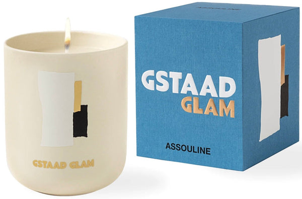 The Travel Candle Gstaad Glam by Assouline is lit in a cream holder with an abstract design beside a blue box, setting the perfect winter escape ambiance.