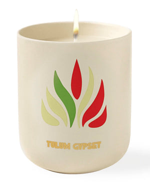 A lit scented candle with an Assouline "Travel Candle Tulum Gypset" logo printed on the side, exuding earthy aromas.