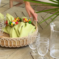A hand arranges a basket with green napkins and beaded napkin rings on the Juliska Provence Rattan Circle Tray atop a wooden table, next to stacked plastic glasses and a clear pitcher.