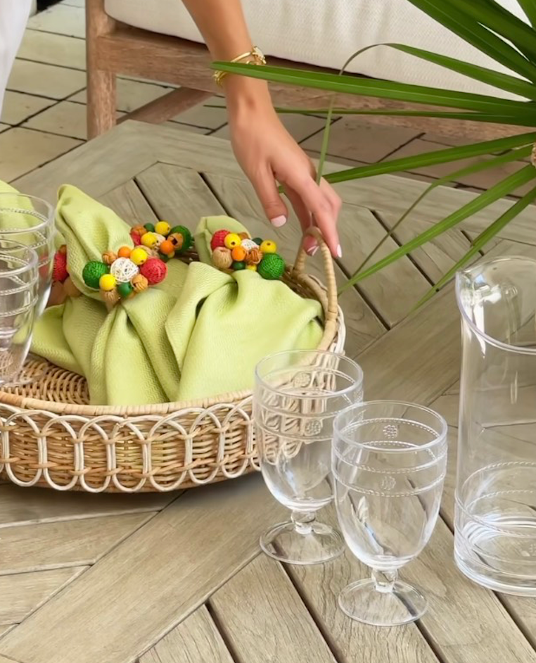 A hand arranges a basket with green napkins and beaded napkin rings on the Juliska Provence Rattan Circle Tray atop a wooden table, next to stacked plastic glasses and a clear pitcher.