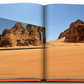 An open book titled "AlUla Ever" by Assouline displays a photograph of a mirrored building reflecting the rocky desert terrain of AlUla, Saudi Arabia, with large sandstone formations under a clear blue sky.
