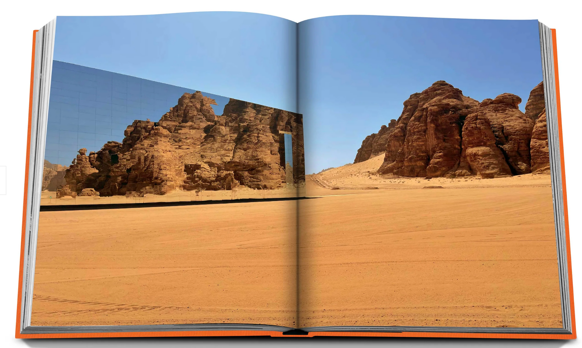 An open book titled "AlUla Ever" by Assouline displays a photograph of a mirrored building reflecting the rocky desert terrain of AlUla, Saudi Arabia, with large sandstone formations under a clear blue sky.