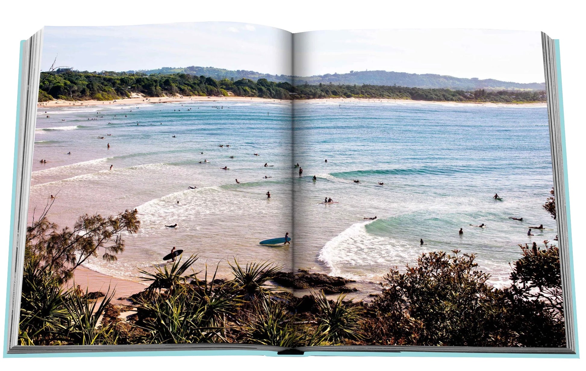 A glimpse into the Byron Bay book by Assouline unveils a lively beach scene, capturing surfers navigating the waves amidst lush greenery and clear skies, embodying the adventurous essence of our Travel Series.