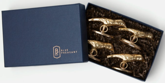 The Hailey Gold Place Card Holder Set by Blue Pheasant features 4 brass bird head holders in a gold finish, elegantly packaged in a dark blue Blue Pheasant box with crinkle paper.