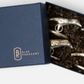 A blue-lidded box with "Blue Pheasant" text and logo contains four Zachary tarnished silver bird-shaped place card holders, nestled in brown shredded paper.