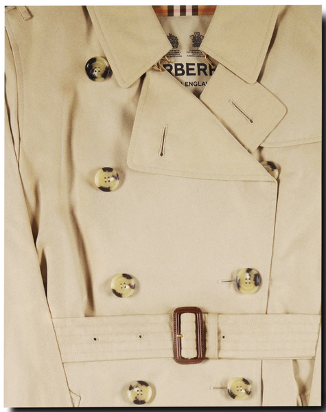 Close-up of a beige double-breasted trench coat with a belt and large buttons, exuding classic British style. A partial label hints at its prestigious origins as featured in "Burberry Book" by Assouline.