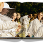 A group of people dressed in white beekeeping suits, some with veiled hats, are gathered together outdoors, engaging in conversation and smiling, reminiscent of Assouline's celebrated Bee Bottle collection featuring Guerlain An Imperial Icon.