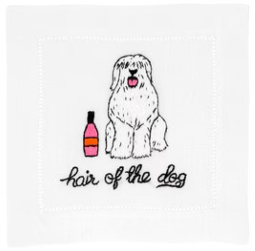 A white dog with long fur sits beside a pink bottle, resting on an elegant cotton napkin by August Morgan. Below, text reads "Hair Of The Dog Cocktail Napkins.