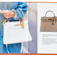 A person in a denim jacket holds a white handbag adorned with a scarf, embodying the essence of luxurious accessories. An open book titled "Little Book of Hermes" by Graphic Image reveals a photo of a brown Hermès handbag on one page, capturing the keen eye of any fashion historian.