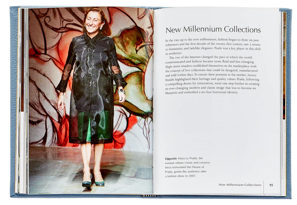 A woman stands smiling in a hallway, wearing a dark Prada dress and sandals, next to a page of the Little Book of Prada by Graphic Image describing the "new millennium collections.