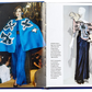 An open "Little Book of Yves Saint Laurent" by Graphic Image displays a model in a blue cape outfit on the left and a mannequin in a similar dress on the right, exploring haute couture designs inspired by the iconic fashion house.