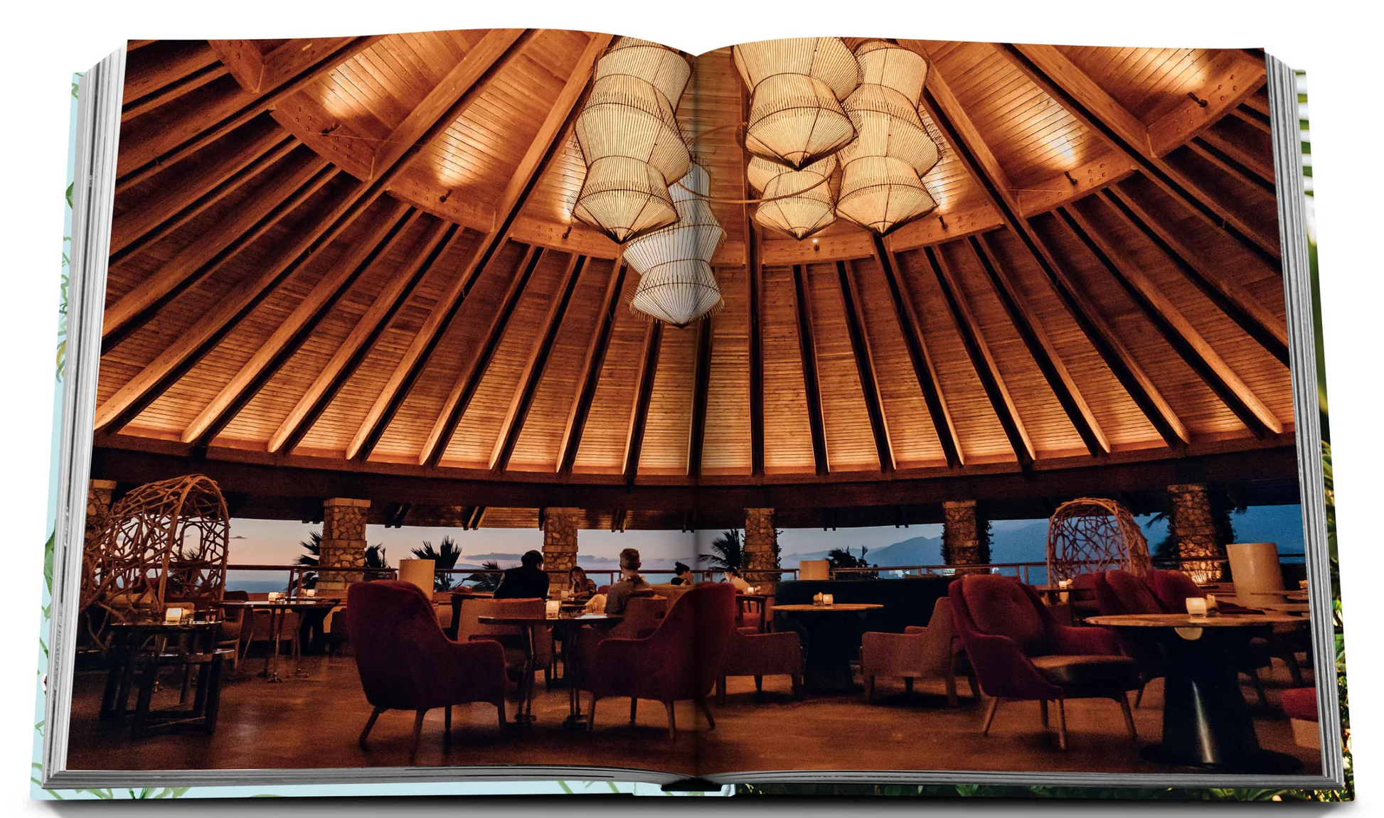 An open book from Assouline's "Hotel Wailea" reveals a restaurant with a round wooden ceiling, warm lighting, various seating arrangements, and an ocean sunset view. This Hawaiian-island escape evokes the serene ambiance of Maui's Hotel Wailea boutique resort.