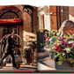 A woman holding a large bouquet of flowers walks past a doorman and bellman at the entrance of Plaza Athénée, renowned for its exquisite French hospitality. The bellman, reminiscent of Jean Imbert's refined elegance, is pushing a luggage cart adorned with "Plaza Athenee" by Assouline.
