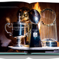 An open book titled "Tiffany & Co.: Crafting Victory" by Assouline showcases three sterling silver sports trophies—a tennis cup, an NFL Super Bowl trophy, and a World Series baseball trophy—set against a dark, dramatic background.