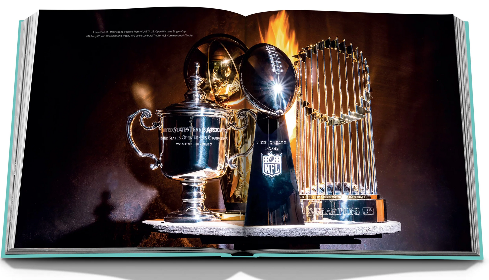 An open book titled "Tiffany & Co.: Crafting Victory" by Assouline showcases three sterling silver sports trophies—a tennis cup, an NFL Super Bowl trophy, and a World Series baseball trophy—set against a dark, dramatic background.