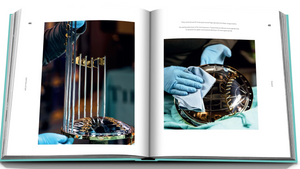 A spread from "Tiffany & Co.: Crafting Victory" by Assouline showcases a silversmithing scene, where gloved hands meticulously polish a trophy, capturing the essence of Tiffany hollowware craftsmanship in sterling silver and its base.