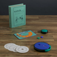Components of the Catch Phrase Vintage Bookshelf Edition by WS Game Company, including a book-style case, game board, circular timer, instruction disks, and colored tokens, are arranged on a wooden surface.
