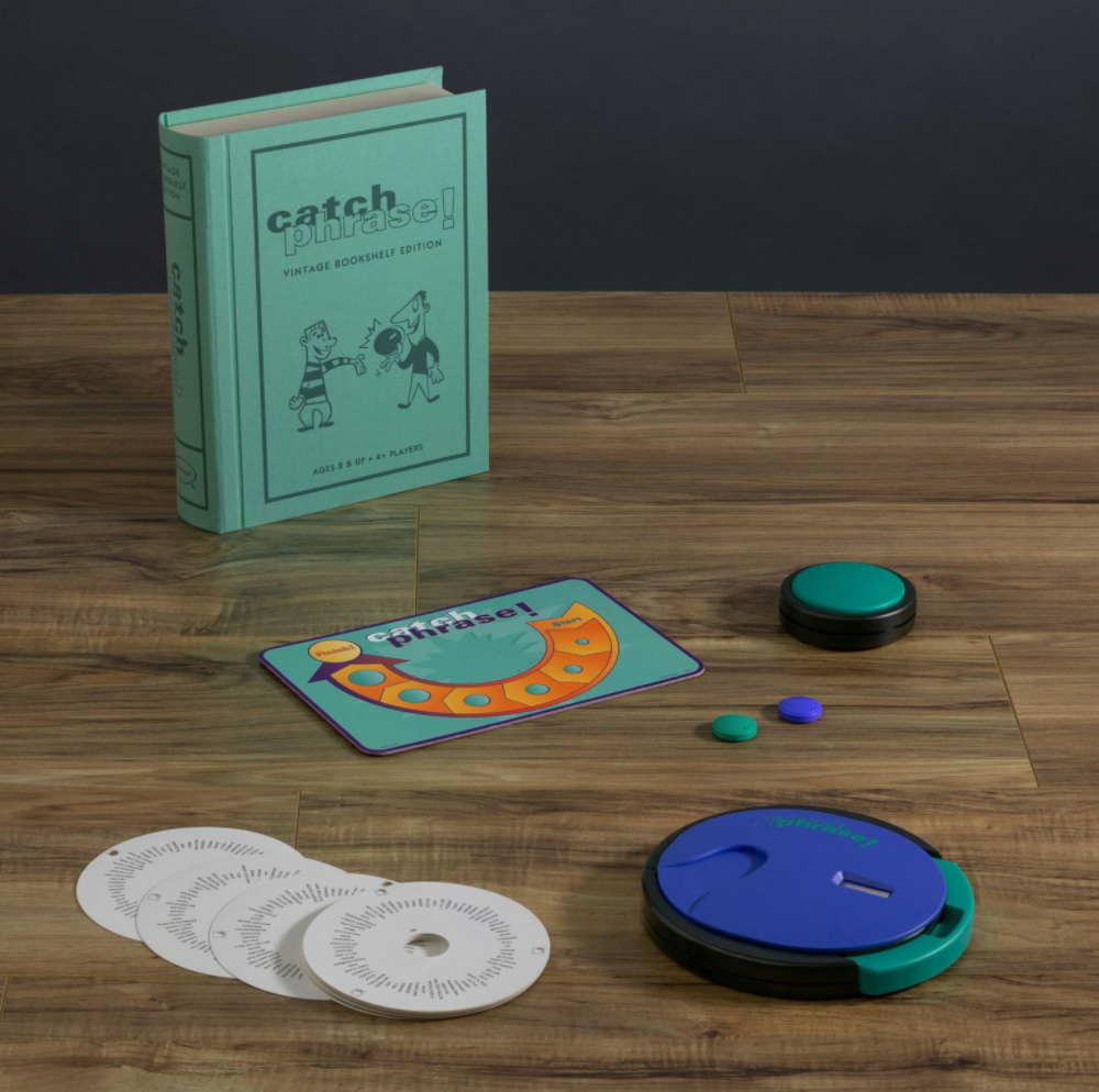 Components of the Catch Phrase Vintage Bookshelf Edition by WS Game Company, including a book-style case, game board, circular timer, instruction disks, and colored tokens, are arranged on a wooden surface.