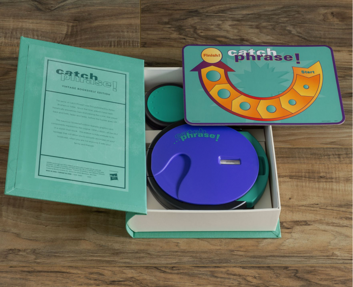 A WS Game Company Vintage Bookshelf Edition of Catch Phrase! featuring a paper insert, timer disk, and game pieces, all neatly arranged in an open box on a wooden surface.