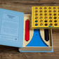An opened Connect Four Vintage Bookshelf Edition set by WS Game Company on a wooden surface. The set features a yellow grid, red and black playing discs, a blue stand, and an instruction booklet nestled in a classic blue box. This vertical strategy game is perfect for adding excitement to game night.
