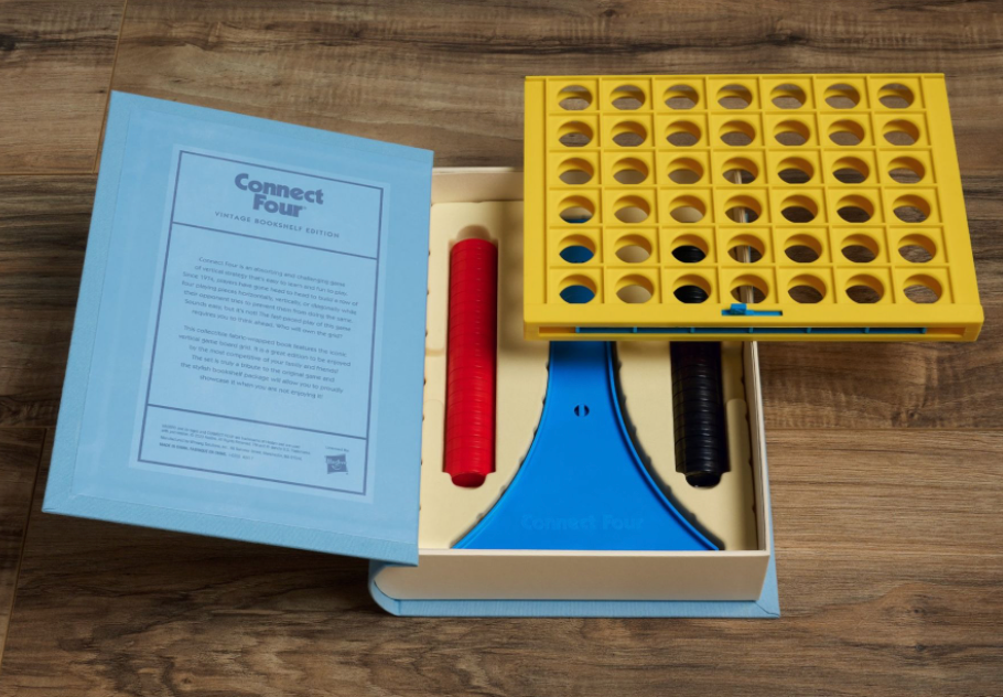 An opened Connect Four Vintage Bookshelf Edition set by WS Game Company on a wooden surface. The set features a yellow grid, red and black playing discs, a blue stand, and an instruction booklet nestled in a classic blue box. This vertical strategy game is perfect for adding excitement to game night.