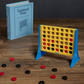 A Connect Four game setup featuring a vertical yellow grid and scattered black and red checkers on a wooden surface, accompanied by a blue book box labeled "Connect Four Vintage Bookshelf Edition" by WS Game Company.