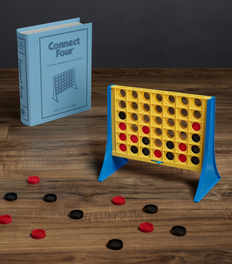 A Connect Four game setup featuring a vertical yellow grid and scattered black and red checkers on a wooden surface, accompanied by a blue book box labeled "Connect Four Vintage Bookshelf Edition" by WS Game Company.