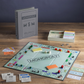 A Monopoly Vintage Bookshelf Edition board game, part of the WS Game Company collection, is displayed on a wooden table. The game board is laid out with cards, money, tokens, dice, and a rule book. The packaging box stands upright beside the game.
