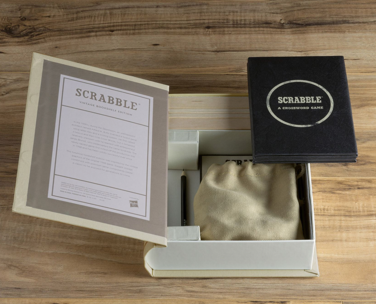 An open box of the Scrabble Vintage Bookshelf Edition by WS Game Company reveals a cloth bag, a pen, and various game components, including a vintage 1948 game board cover and an instruction sheet, all elegantly displayed on a wooden surface adorned with classic wood letter tiles.