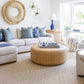 A bright living room features a white sectional sofa, blue and green patterned pillows, and the Moselle Coffee Table by Made Goods—a large, round rattan drum—placed by the window to let in natural light.