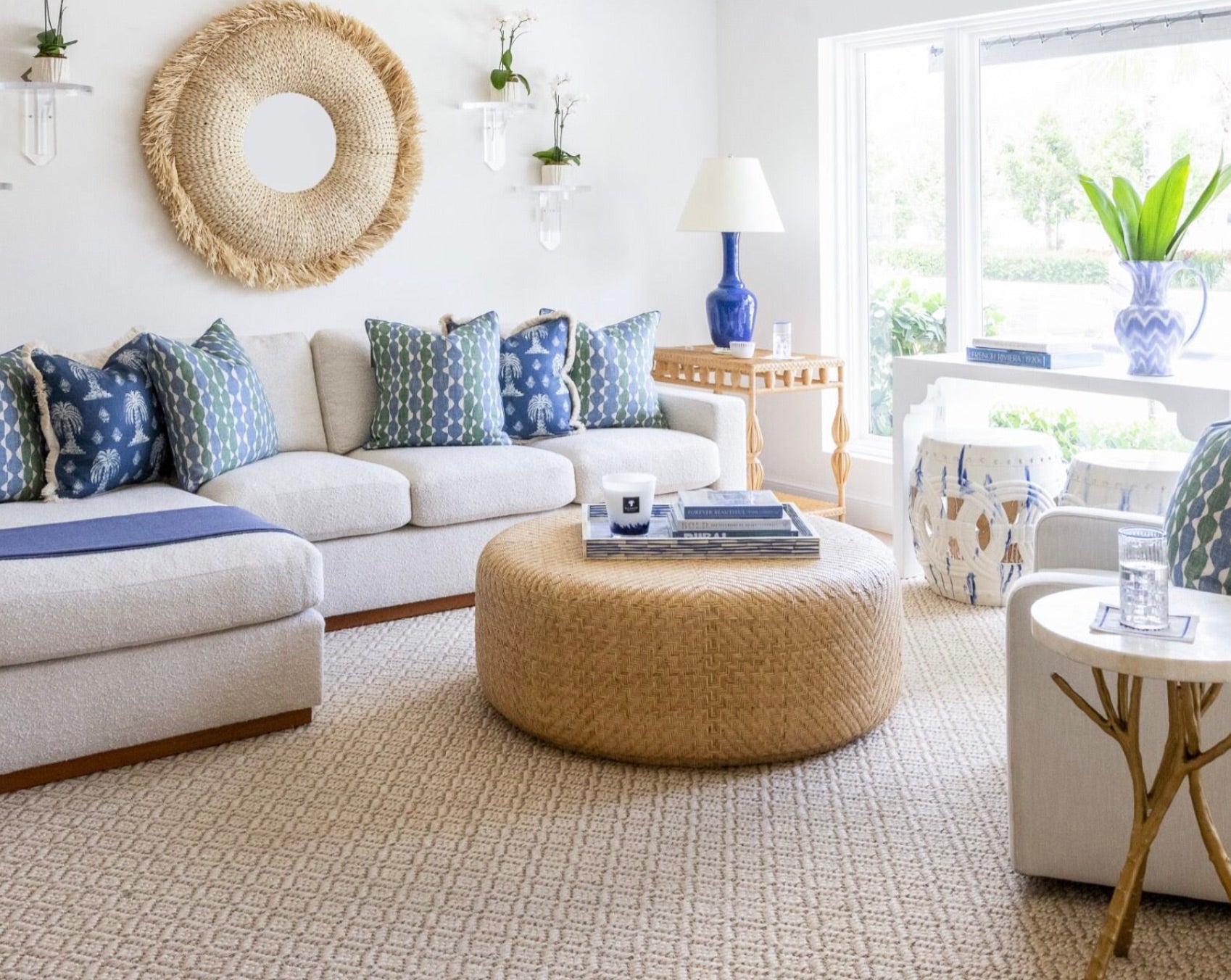 A bright living room features a white sectional sofa, blue and green patterned pillows, and the Moselle Coffee Table by Made Goods—a large, round rattan drum—placed by the window to let in natural light.