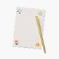 A gold pen rests on a Riffle Paper Co. Dahlia Collection notepad with scalloped edges and small corner floral designs, set against a light background.