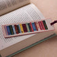 The Rifle Paper Co. Bookmark Collection features a fabric bookmark with colorful embroidered book spines and whimsical print, shown resting on an open book atop a beige surface.
