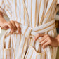 A person ties the belt of a Weezie Towels Short Plush Robe, holding the soft striped fabric near the waist.