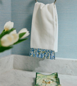 The Weezie Towels x Liberty London Powder Bath Towel, a white terry towel trimmed with blue Liberty London fabric, hangs on a silver ring above a marble countertop—ideal for adding style and function to your powder bath.