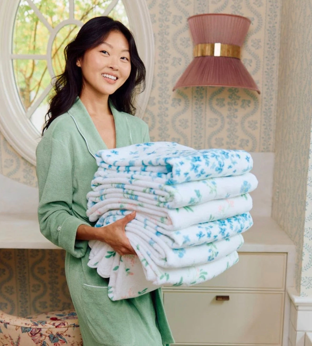 A woman in a green robe holds a large stack of Weezie Towels' Betty Floral Scallop Towel Collection in a room with patterned wallpaper and a mirror.