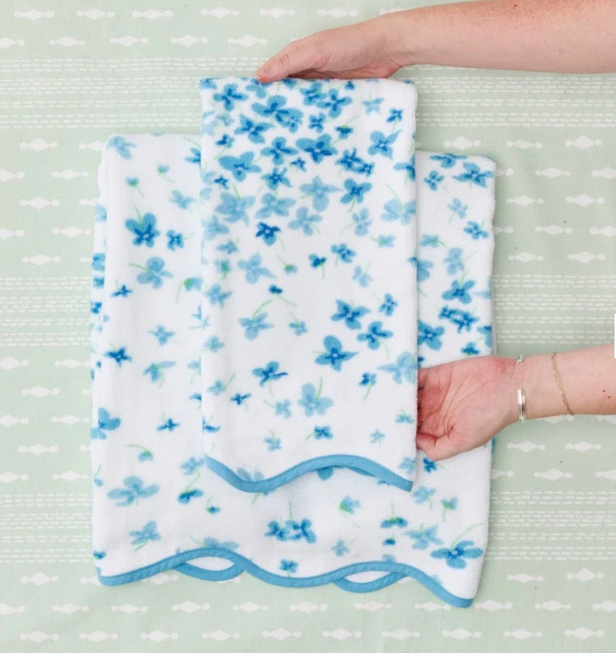 Two hands hold small and medium towels from the Betty Floral Scallop Towel Collection by Weezie Towels, placed on a larger matching towel atop a pale green patterned surface—an elegant addition to your bathroom décor.