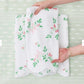Two hands arrange a white towel from the Alice Floral Scallop Towel Collection by Weezie Towels, featuring a pink and green floral pattern and hand-finished scallop trim, on a light green patterned surface.